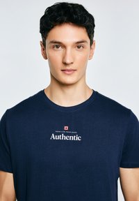 Jonge man met kort zwart haar, gekleed in een donkerblauw T-shirt met de tekst "Authentic" op de voorkant, tegen een effen lichte achtergrond.