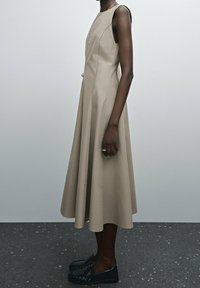 Beige A-linjainen mekko, jossa on hihaton muotoilu, avonainen selkä ja sauman yksityiskohtia. Yhdistetty mustiin liukupohjiin.