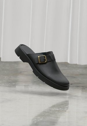 Zoccolo slip-on in pelle nera con suola spessa e una grande fibbia dorata sulla parte superiore, posizionato su un pavimento riflettente con sfondo di tende bianche.