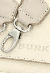 Detail einer hellbeigen Lederhandtasche mit silberfarbener Hardware und genähten Akzenten, auf deren Oberfläche "BURK" geprägt ist.