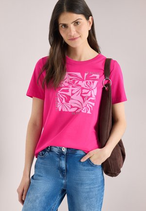 Jeune femme aux longs cheveux noirs portant un t-shirt graphique rose vif, un jean bleu, et un sac à bandoulière marron foncé.