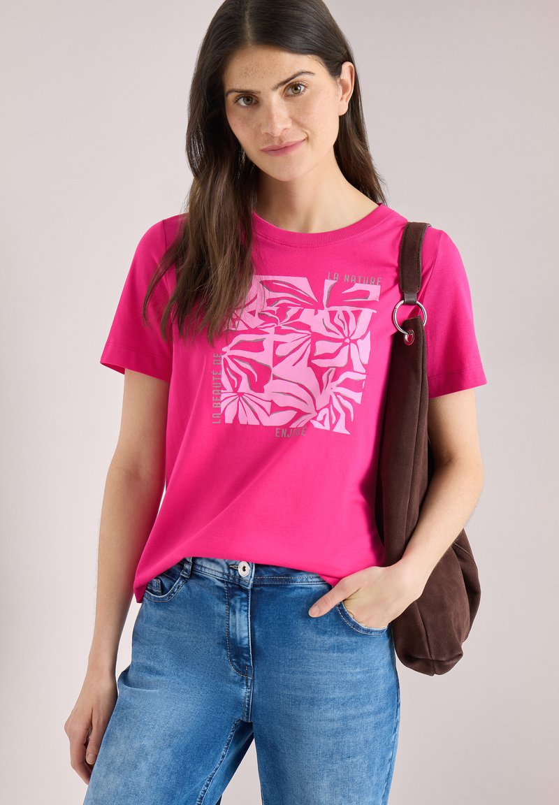 Joven con cabello largo y oscuro, lleva una camiseta gráfica de color rosa brillante, vaqueros azules y lleva un bolso de hombro marrón oscuro.