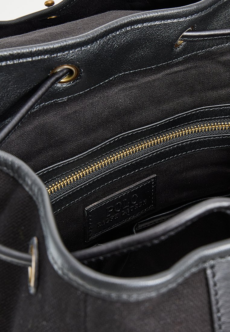 Schwarze Tragetasche mit strukturiertem Äußeren, Lederakzenten und einem goldenen Reißverschlussfach innen. Mit einem Textilfutter und einem Logo-Tag ausgestattet.