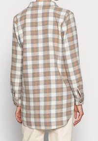 Achteraanzicht van een persoon in een langslavend, beige en blauw geruite blouse met knopen aan de manchetten en crèmekleurige broek.