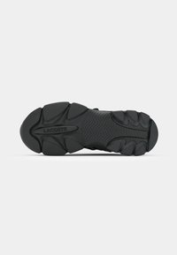 Semelle en caoutchouc noir d’une chaussure Lacoste avec des motifs de bande de roulement texturés et le nom de la marque « LACOSTE » embossé près du talon et des orteils.