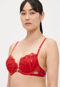 Raudonos nėriniuotos liemenėlės, kurių konstrukcija yra struktūruota su geometriniais raštais, satino akcentais, reguliuojamomis petnešėlėmis ir centrinio dekoro sagtimi.