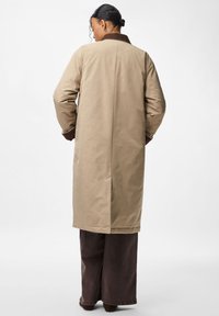 Casaco longo bege com gola castanha, apresentando um design direito e ajuste descontraído. Costuras visíveis ao longo das costuras e punhos.