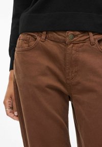 Bruine katoenen broek met een klassieke vijf-pocketstijl, zichtbaar stikwerk en een metalen knoop in de taille.