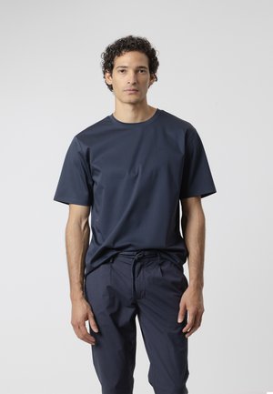 Hombre con cabello rizado y oscuro que lleva una camiseta azul marino lisa y pantalones a juego con cordón, de pie frente a un fondo gris claro.