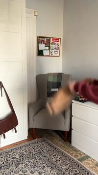 Szürke kárpitozott fotel, kúpos fából készült lábakkal, a háttámláján egy kockás gyapjú takaró, és egy közeli barna bőrtáska.