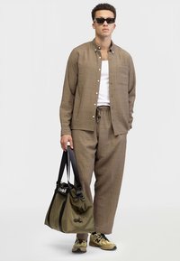 Camicia marrone chiaro con texture, stile abbottonato con taschino sul petto, abbinata a pantaloni larghi dello stesso colore. La modella tiene una borsa verde oliva scuro con manici neri.