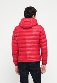 Man wearing a bright red quilted puffer jacket with a hood, paired with dark blue jeans, standing with back facing viewer.