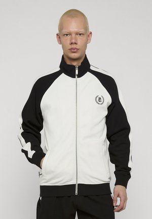 Młoda osoba o krótkich blond włosach, ubrana w białą i czarną kurtkę na zamek z małym herbem na piersi, stojąca z rękami w kieszeniach.