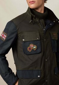 Muškarac u maslinasto zelenoj i mornarsko plavoj jakni s oznakama Union Jack i Hackett London na rukavu i džepovima na prsima, ispod nosi crnu rolku.