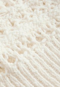 Gros plan sur un tissu tricoté doux et blanc montrant des motifs détaillés et texturés avec une combinaison de côtes et de points de dentelle.