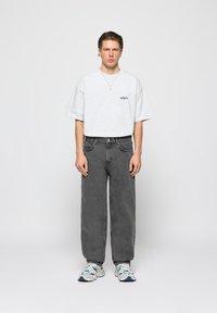 Młody mężczyzna stoi, ubrany w białą, oversize'ową koszulkę, luźne ciemnoszare dżinsy, białe trampki i złoty naszyjnik na gładkim jasnym tle.