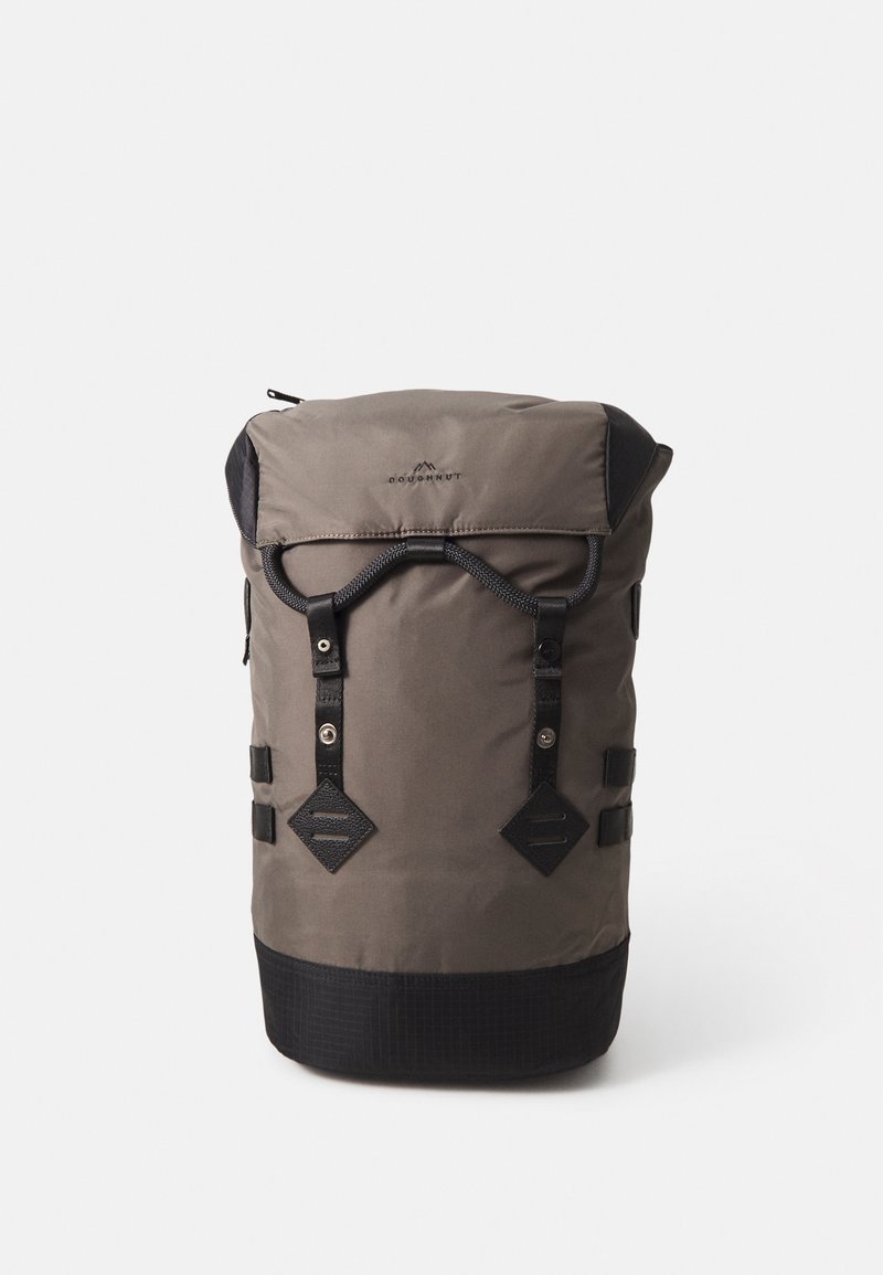Sac à dos en tissu gris avec un design à rabat, accents noirs, deux sangles latérales et quincaillerie en forme de diamant à l'avant. Fond texturé.