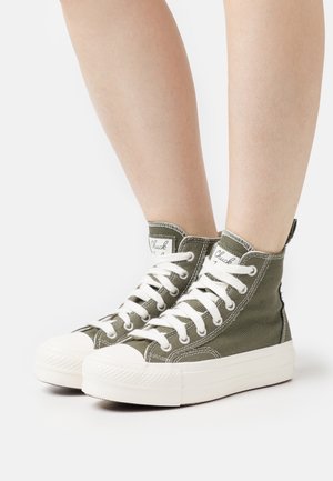 Beine in olivgrünen High-Top-Canvas-Sneakers mit weißen Gummisohlen und weißen Schnürsenkeln auf weißem Hintergrund.