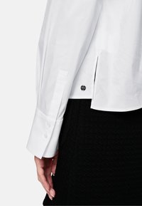 White button-up shirt featuring a smooth texture, long sleeves with button cuffs, and side slits. Worn over a black textured skirt.