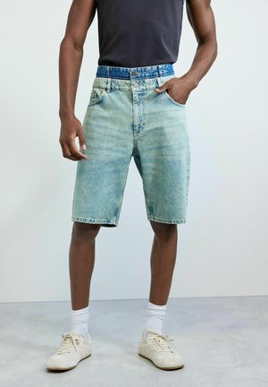 Férfi világoskék farmer rövidnadrágban, kettős gombos derékrésszel, fekete ujjatlan pólóban, fehér zokniban és bézs sportcipőben, zsebében tartott kézzel áll.