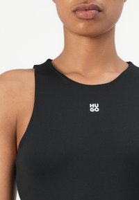 Černý bezrukávový body z hladkého materiálu, s bílým logem "HUGO" na střední části hrudi a kulatým výstřihem.