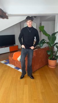 Jeune homme en pull noir et jean debout dans un salon avec un canapé orange, une grande plante verte et un meuble TV en arrière-plan.