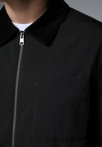 Chaqueta negra con cuello texturizado, cremallera frontal y bolsillo en el pecho. El material parece suave con un sutil detalle de costura.