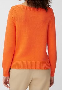 Persona che indossa un vivace maglione di lana arancione con polsini e orlo a coste, abbinato a pantaloni beige, visto da dietro.