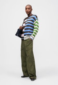 Pulover tricotat cu dungi orizontale multicolore în nuanțe de albastru, marron, verde și roz. Pantaloni cargo olive cu buzunare laterale și pantofi negri.