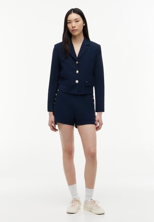 Kvinde iført en marineblå blazer med guldknapper og matchende højtaljede shorts, hvide sokker og beige sneakers, stående foran en hvid baggrund.