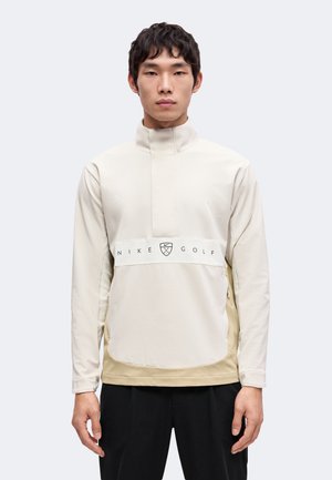 Junger Mann trägt einen beigen Nike Golf Pullover mit hohem Kragen, steht vor einem schlichten hellen Hintergrund und blickt nach vorne.
