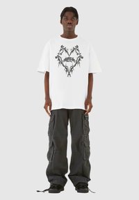 Hvit oversized t-skjorte med et svart blomster hjerte grafisk design, paret med svarte cargo bukser med flere lommer og snøringdetaljer.