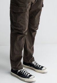 Bruine cargobroek met zijzakken; gemaakt van een lichtgewicht stof. Gepaard met zwarte high-top sneakers met witte rubberen zolen.