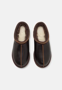 Brown leather slippers with a soft lining, rounded toe, and contrast stitching around the edge, featuring a subtle logo on the side.