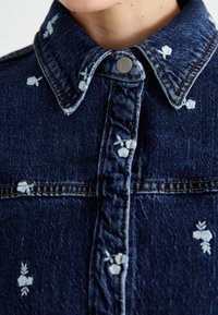 Casaco de ganga azul escuro com gola, apresentando bordados florais brancos e costuras amarelas em contraste ao longo das costuras.