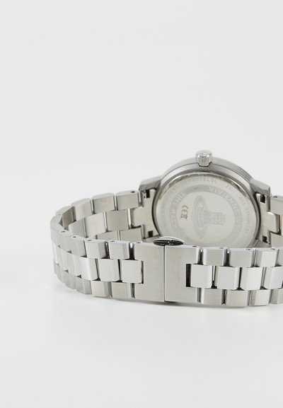 Relógio em aço inoxidável prateado com caixa redonda, pulseira texturizada metálica e fecho de pressão. A parte de trás apresenta uma gravação detalhada.