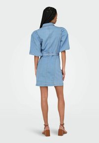 Denimmekko, jossa on lyhyet puff-hihat, vyötärönauha, takasaumadetaljointi ja vaaleansininen väri. Asusteena ruskeat paksupohjaiset sandaalit.