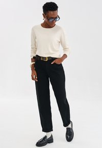 Jasno beżowy sweter z długim rękawem, czarne spodnie o wysokim stanie z podwiniętymi mankietami, brązowy pasek z złotą klamrą oraz błyszczące czarne mokasyny.