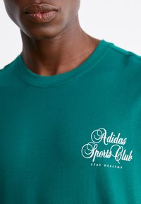 Turkise bomulds t-shirt med rund halsudskæring og korte ærmer, pyntet med hvid tekst, der står "Adidas Sports Club" og "STAY HEALTHY."