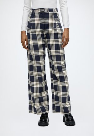Persona che indossa pantaloni a vita alta a gamba larga a quadri blu navy e crema, abbinati a mocassini neri chunky e una maglietta bianca a maniche lunghe.