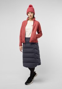 Veste en polaire rouge avec un col haut, t-shirt crème, jupe rembourrée noire, et baskets noires. Bonnet en tricot avec détail en pompon, en tissu texturé.