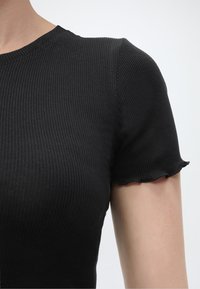 Κοντινό πλάνο ενός ατόμου που φοράει μαύρο ριμπ κοντομάνικο μπλουζάκι με διακριτικές κυματιστές άκρες στα μανίκια, πάνω σε απλό φωτεινό φόντο.