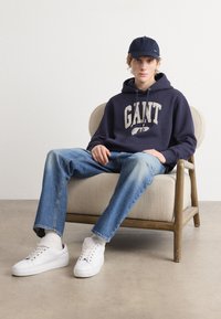 Marineblå hettegenser med "GANT" trykk, lyse denimjeans og hvite joggesko. Modell som sitter i en beige polstret stol mot en ensfarget bakgrunn.