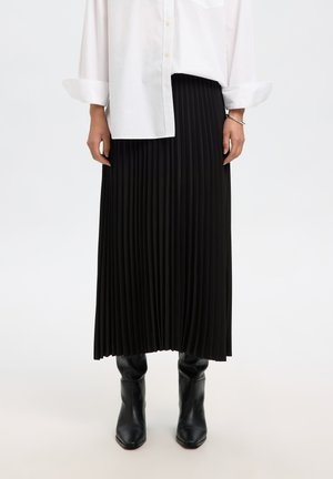 Femme portant une chemise blanche boutonnée, une jupe midi plissée noire, un bracelet argenté et des bottes en cuir noires, debout sur un fond uni.