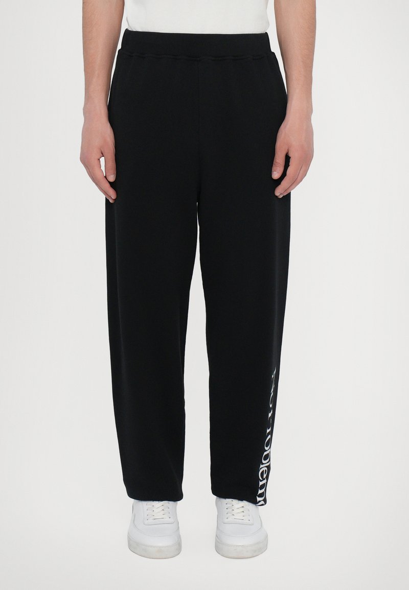 Black sweatpants with an elastic waistband and a loose fit. Notable detail includes white text printed along the left leg.