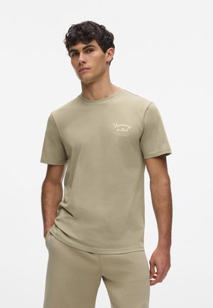 Jeune homme aux cheveux bouclés foncés portant un t-shirt beige et un pantalon assorti, debout avec une main dans la poche sur fond blanc.