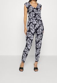 Woman wearing sleeveless black jumpsuit with white floral print, belted waist, and black slide sandals, standing against white background.