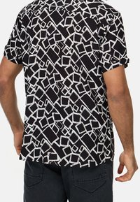 Zwart shirt met korte mouwen, voorzien van een wit geometrisch patroon van overlappende vormen en lijnen, gemaakt van lichtgewicht stof.