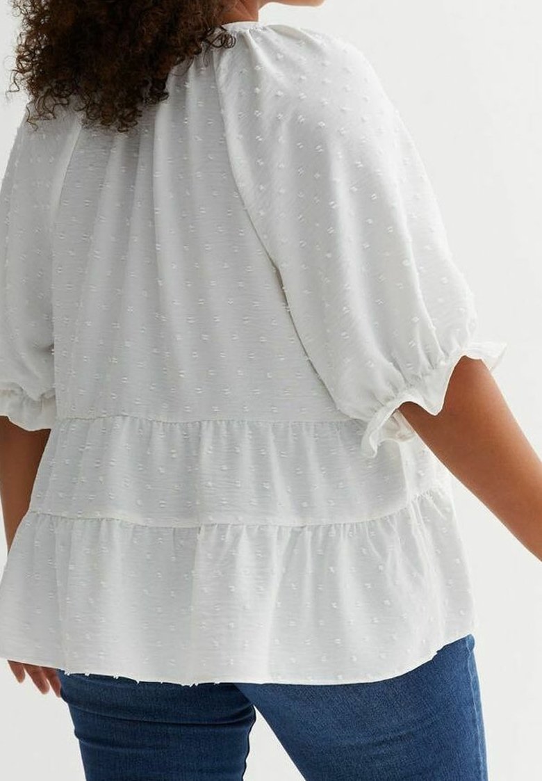 Žena v bílé texturované blůze s puffovými rukávy a volány, která je spárovaná s tmavě modrými džínami, zobrazená zezadu.