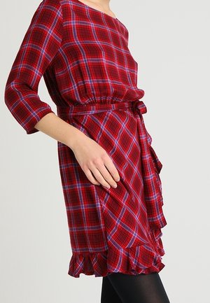 Rotes kariertes Kleid mit dreiviertellangen Ärmeln, tailliertem Gürtel, gerafftem Saum und einer Hand, die sich an der Hüfte gegen einen einfarbigen Hintergrund abstützt.